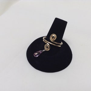 May include: A gold wire ring with a pink teardrop-shaped gemstone dangling from it. The ring is displayed on a black velvet ring holder.