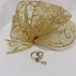 May include: A gold-toned ring with a spiral design and a dangling pink gemstone. The ring is on a white surface with a gold and white ribbon in the background.