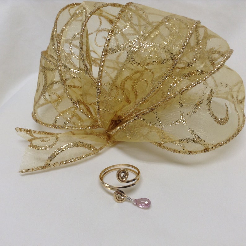 May include: A gold-toned ring with a spiral design and a dangling pink gemstone. The ring is on a white surface with a gold and white ribbon in the background.