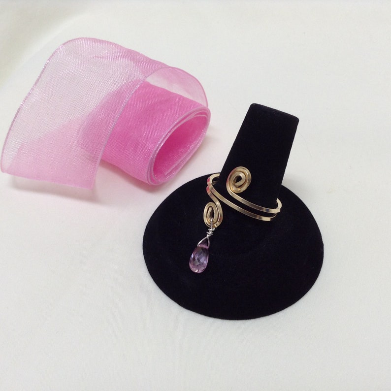 May include: A gold ring with a spiral design and a dangling pink gemstone. The ring is displayed on a black ring holder.