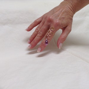 May include: A gold wire ring with a dangling purple gemstone. The ring is worn on the index finger of a hand.