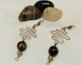 Peut inclure: Boucles d'oreilles dorées avec un design de fil ondulé et une perle en œil de tigre marron.