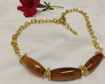 Peut inclure: Un collier à chaîne dorée avec trois grandes perles marron en forme de tonneau. Les perles présentent un aspect craquelé et à motifs. Une fleur rouge est en arrière-plan. Ce collier est un accessoire de mode.
