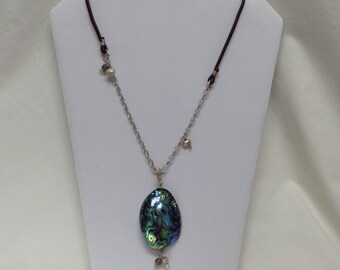 Peut inclure: Un collier en chaîne argentée avec un cordon en cuir marron et un grand pendentif en nacre d'ormeau iridescent. Le pendentif est entouré de petites perles blanches et grises.