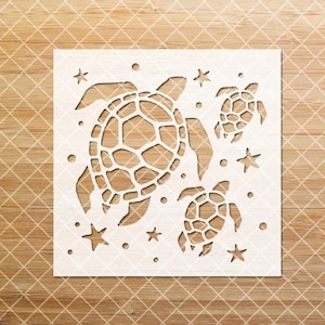 May include: A white stencil featuring three sea turtles of varying sizes, surrounded by stars and small circles. The stencil is cut from a square piece of material and is set against a light brown, wood-grain background.