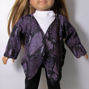 May include: A doll wearing a purple and black snakeskin-patterned jacket over a white turtleneck and black pants. The doll has long, light brown hair and green eyes. The jacket has long sleeves and a V-neck design.