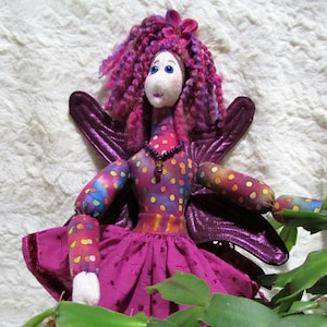 May include: A handmade purple fairy doll with pink yarn hair, a purple and white polka dot dress, and iridescent purple wings. The doll is posed with its arms outstretched and is sitting in a green plant.