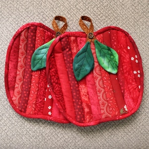 May include: Two apple-shaped fabric potholders constructed from vertical strips of red patterned cotton fabric. Each potholder features two green fabric leaves and a brown fabric loop attached with a small brown button at the top center.