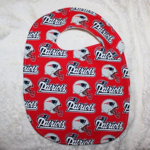 May include: A red baby bib featuring a repeating pattern of the New England Patriots football team logo, which consists of a silver helmet with a blue and red face mask and the word Patriots written in a bold, white, stylized font.