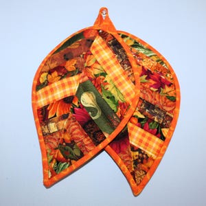 May include: A pair of orange and brown quilted pot holders shaped like leaves with a fall-themed fabric design.