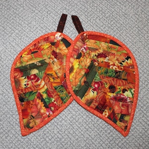 May include: Two leaf-shaped quilted pot holders featuring a patchwork design of autumn-themed fabric with sunflowers, pumpkins, and fall foliage in shades of orange, red, yellow, and green. Each pot holder has an orange border and a brown fabric loop.