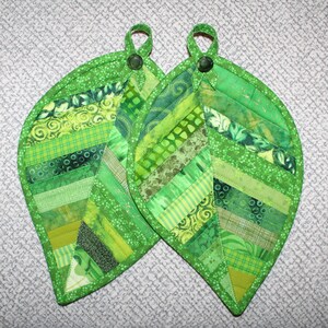 May include: Two green leaf-shaped pot holders made from patchwork fabric in various shades of green. The pot holders have a button on the top.