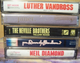 May include: A stack of five cassette tapes. The top tape is white with red text that reads "Luther Vandross Any Love". The second tape is purple with white text that reads "Janet.". The third tape is black with yellow text that reads "The Neville Brothers Yellow Moon". The fourth tape is blue with white text that reads "Pam Dawn The Bliss Album". The bottom tape is white with red text that reads "Neil Diamond 12 Greatest Hits Vol II".