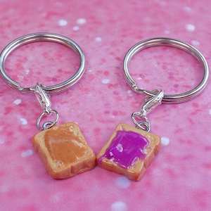 May include: Two keychains with miniature toast slices. One has peanut butter, the other jelly. Each keychain has a silver ring and clasp. The background is a pink surface with white speckles.