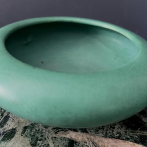 May include: A low-profile, circular bowl with a smooth, matte finish in a muted seafoam green colour. The vessel features a wide, rounded rim that curves inward towards a deep, open centre, creating a simple, continuous, and rounded silhouette.