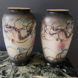 May include: A pair of ceramic vases featuring a gradient cream and black finish with raised relief dragon motifs in gold, brown, and light blue. The vases have a narrow neck with a gold rim and are decorated with swirling black line patterns.