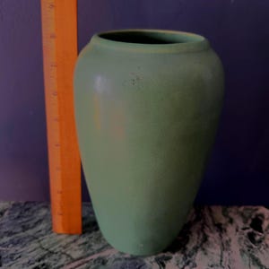 Puede incluir: Un jarrón de color verde salvia mate con cuerpo redondeado y una abertura ancha. Una regla naranja está colocada verticalmente junto al jarrón, mostrando una altura de aproximadamente 23 cm. El jarrón tiene una superficie lisa y una forma simple.