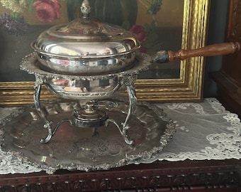 Può includere: Un chafing dish argentato con coperchio e manico in legno. Il piatto poggia su un supporto decorativo con gambe ornate e un vassoio da portata abbinato. Il chafing dish è progettato per servire cibo.