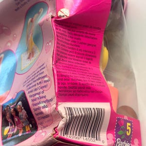 May include: A pink Barbie product packaging box featuring a barcode, a small yellow warning icon for children under 36 months, and multi-language text including 'Collect all her Lil' Friends!' and 'Barbie' with a number 5 logo.