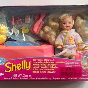 May include: A Shelly doll set in original packaging featuring a blonde doll in a white bathrobe with yellow duck print, a pink bathtub, a yellow rubber duck, a pink comb, and various small bath accessories. The box text reads Bathtime Fun Shelly.