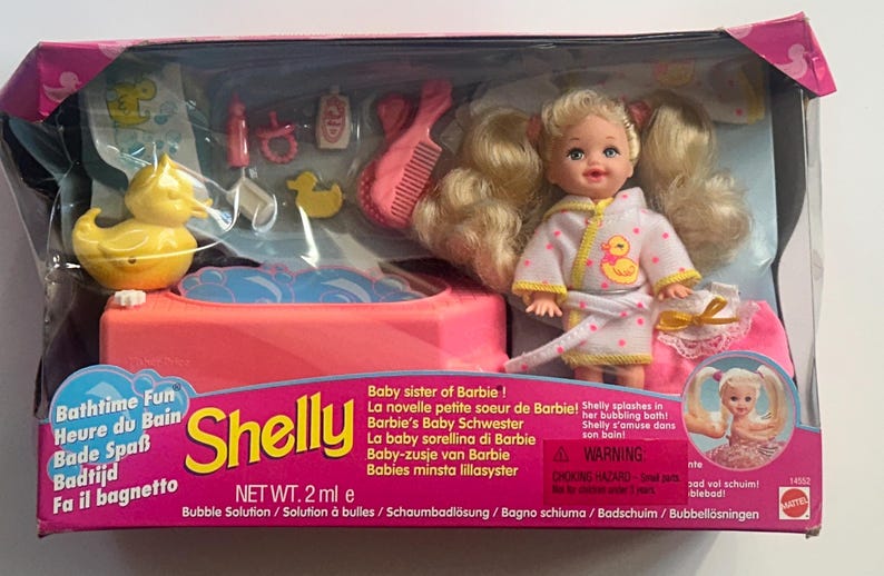 May include: A Shelly doll set in original packaging featuring a blonde doll in a white bathrobe with yellow duck print, a pink bathtub, a yellow rubber duck, a pink comb, and various small bath accessories. The box text reads Bathtime Fun Shelly.
