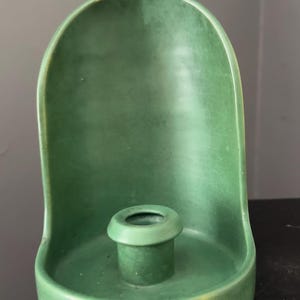 Puede incluir: Un candelabro de cerámica verde mate con un panel trasero redondeado en forma de arco y un soporte cilíndrico central para una vela. La superficie muestra una textura de esmalte craquelado sutil con tenues reflejos amarillos en los bordes.