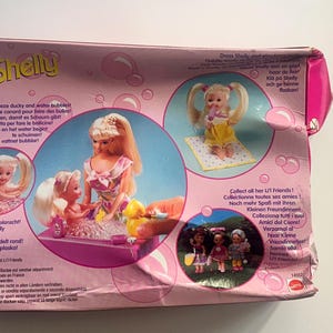 May include: A pink cardboard toy packaging box for a Shelly doll set, featuring illustrations of dolls in a bathtub. A wooden ruler placed to the right shows the box is approximately 23 cm wide. The packaging includes text in multiple languages.