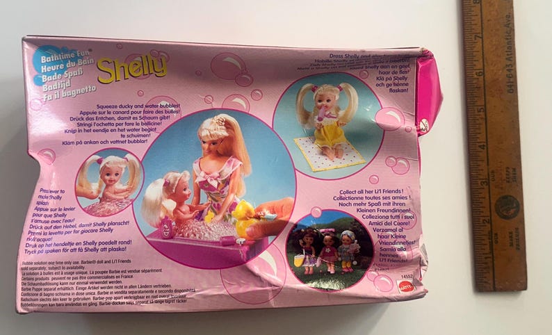 May include: A pink cardboard toy packaging box for a Shelly doll set, featuring illustrations of dolls in a bathtub. A wooden ruler placed to the right shows the box is approximately 23 cm wide. The packaging includes text in multiple languages.