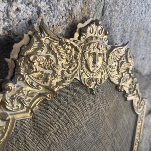 Puede incluir: Una pantalla para chimenea de metal dorado con un borde superior ornamentado que presenta un relieve de un rostro humano central, flanqueado por figuras aladas y patrones de volutas intrincados. El cuerpo principal es una malla metálica fina.
