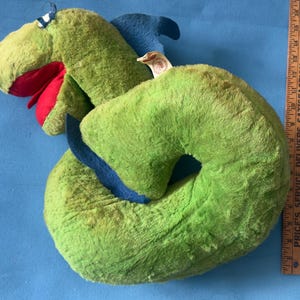 May include: A green plush dragon toy with a red mouth, blue fins, and a white tag, positioned next to a wooden ruler showing a length of approximately 17 inches. The dragon has a curved body and a textured, soft fabric surface throughout its design.
