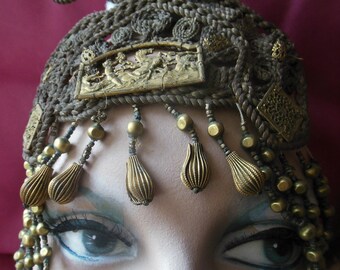 May include: A gold-coloured headpiece with intricate braided designs and dangling gold beads. The headpiece is worn by a person with makeup and blue eyes.
