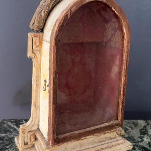 May include: An arched wooden display case with a weathered cream and brown finish. The interior features a deep red fabric backing with a subtle floral pattern. A small metal latch is attached to the side of the frame, and a decorative carved element sits atop the arched structure.