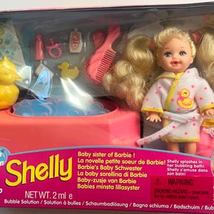 May include: A Shelly doll with blonde hair in pigtails wearing a white bathrobe with pink dots and a yellow duck graphic. The set includes a pink bathtub, a yellow rubber duck, a pink comb, a bottle, and a pacifier in packaging.
