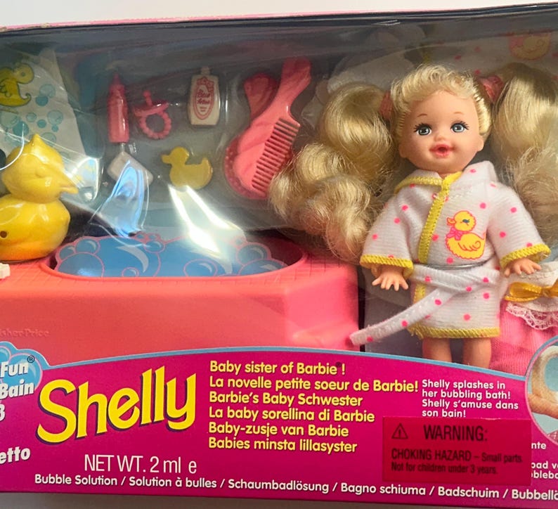 May include: A Shelly doll with blonde hair in pigtails wearing a white bathrobe with pink dots and a yellow duck graphic. The set includes a pink bathtub, a yellow rubber duck, a pink comb, a bottle, and a pacifier in packaging.