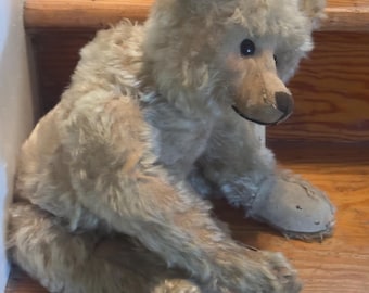 Puede incluir: Un oso de peluche con pelaje beige claro, ojos de color marrón oscuro y una nariz cosida de color marrón oscuro. El oso tiene almohadillas de tela clara en las patas y está sentado de perfil, mostrando su pelaje texturizado y extremidades.