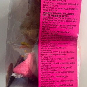 May include: A pink toy packaging box featuring a clear plastic window revealing a doll with blonde hair and a pink skirt. The side panel displays white text detailing Mattel manufacturing information, safety standards, and company addresses.