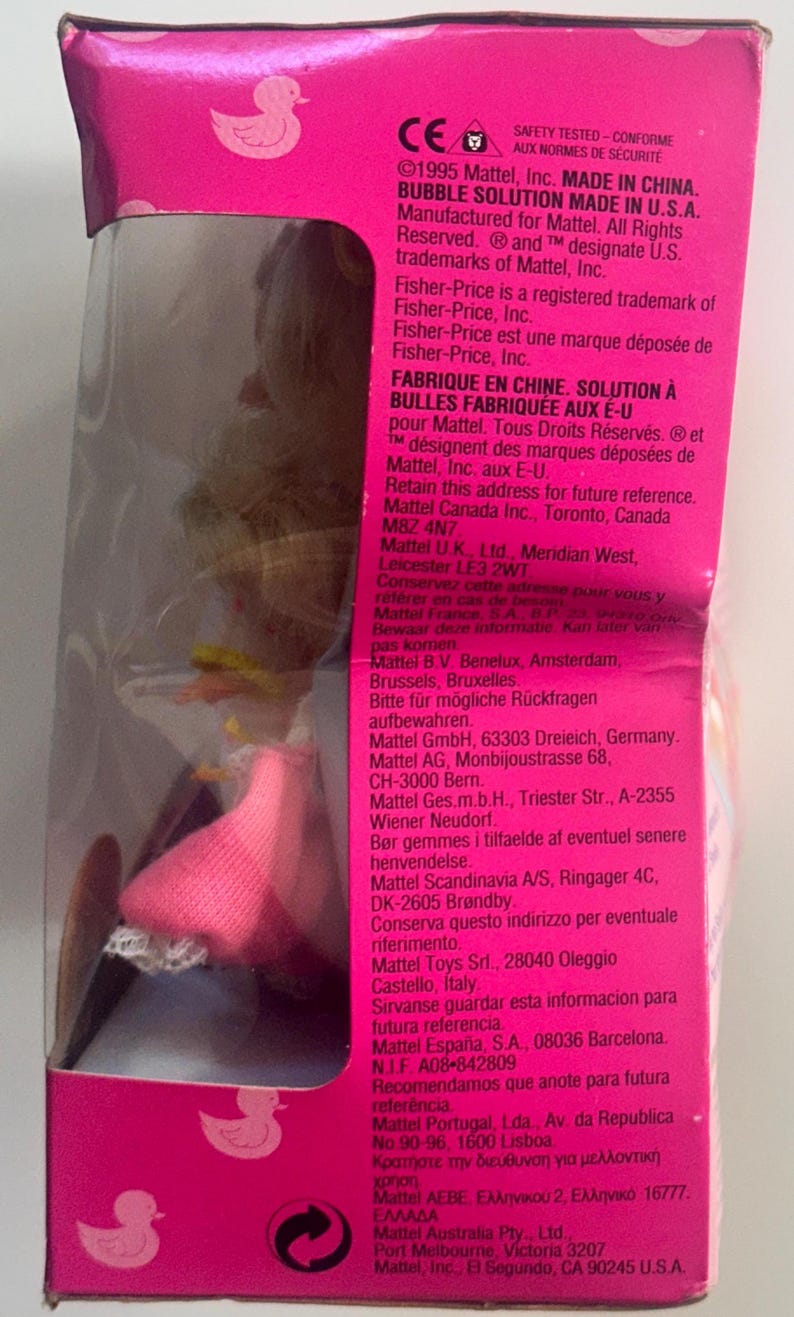 May include: A pink toy packaging box featuring a clear plastic window revealing a doll with blonde hair and a pink skirt. The side panel displays white text detailing Mattel manufacturing information, safety standards, and company addresses.