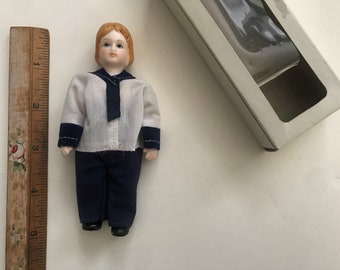 May include: A porcelain doll wearing a white sailor shirt with blue trim and blue trousers. The doll has red hair and blue eyes.