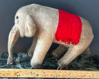 Puede incluir: Un juguete de elefante de peluche gris vintage con una montura roja. El elefante tiene una larga trompa, ojos negros pequeños y está de pie sobre cuatro patas. El juguete probablemente está hecho de fieltro o un material similar.