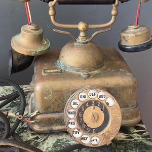 Puede incluir: Teléfono antiguo de latón con auricular y microteléfono negros. El teléfono tiene un dial circular con botones con letras blancas y un cable negro. El teléfono está sobre una superficie de mármol verde y blanco.