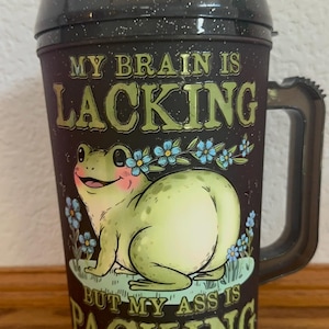 May include: Black travel mug with a humorous frog illustration and the text "MY BRAIN IS LACKING BUT MY ASS IS PACKING." The frog is light green with pink cheeks and blue flower accents. The mug has a black handle and lid.