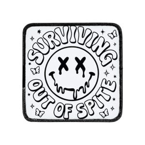 May include: A white square patch with a black border. The patch features a black and white design with the words "SURVIVING OUT OF SPITE" in a circular pattern around a smiley face with X eyes and dripping mouth. Butterflies and stars are also included.