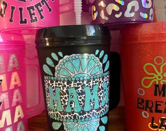 Puede incluir: Vaso negro con asa, con la palabra "MAMA" en turquesa, estampado de leopardo y detalles brillantes. Otros vasos en rosa, morado y rojo con varios diseños son visibles en el fondo.