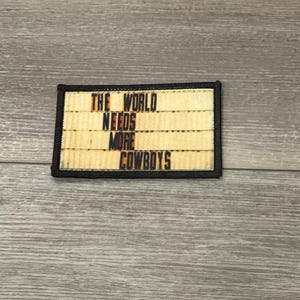 May include: A black and yellow embroidered patch with the text "The World Needs More Cowboys" on a beige background.