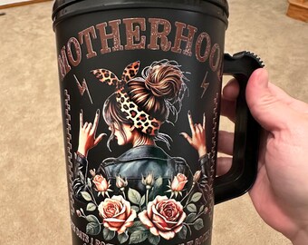 Puede incluir: Taza de viaje negra con asa y pajita. Tiene un gráfico de una mujer con un pañuelo de leopardo, una chaqueta de cuero y rosas. El texto en la taza dice "Motherhood, Some Days I Rock It, Some Days It Rocks, Either Way We're Rockin".