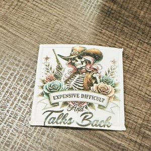 May include: A white patch with a skeleton wearing a cowboy hat and holding a gun. The skeleton is surrounded by flowers. The text on the patch reads "Expensive Difficult And Talks Back".