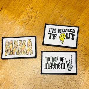 May include: Three iron-on patches with white backgrounds and black borders. The first patch says "MAMA" in floral letters. The second patch says "I'M MOMED TF OUT" with a yellow smiley face. The third patch says "MOTHER OF MAYHEM" with a black skeleton hand making a rock and roll sign.