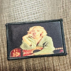 May include: A black and white embroidered patch featuring a woman with blonde hair, wearing a green sweater and holding a guitar. The background is black.