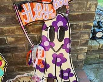 Puede incluir: Un colgador de puerta de fantasma blanco con flores moradas y un contorno negro. El fantasma sostiene una pancarta que dice "Hey Boo" en letras rosas y naranjas. El fantasma tiene ojos negros y una sonrisa negra. El colgador de puerta está decorado con purpurina y tiene una cinta rosa y naranja colgando.