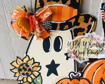 Puede incluir: Colgador de puerta de madera con forma de fantasma con un sombrero de vaquero con diseño de estampado de leopardo. El fantasma tiene ojos negros y una estrella. También se incluye un girasol y una calabaza. El colgador tiene un lazo decorativo con detalles en naranja y negro.